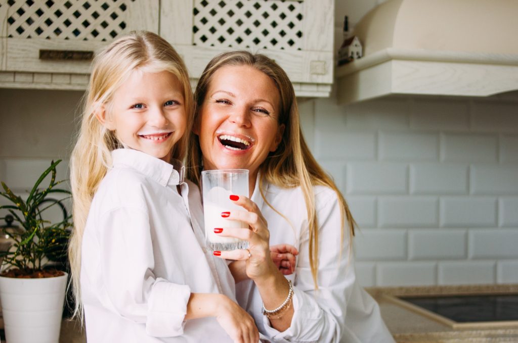 Happy blonde long hair mom and daughter drinking milk in kitchen, healthy lifestyle