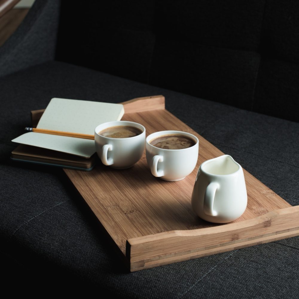 Two cups of coffee, jug of milk and notebook with pencil on wooden tray on sofa
