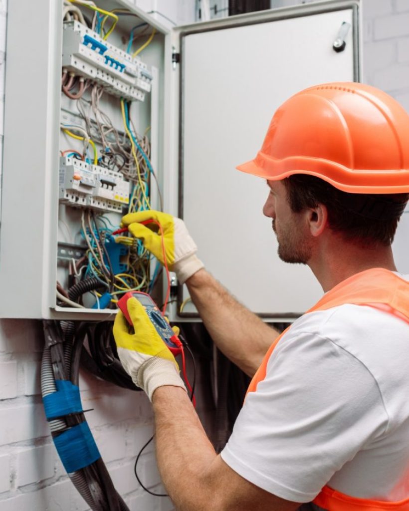 Handsome electrician in hardhat and gloves holding multimeter near electrical distribution box