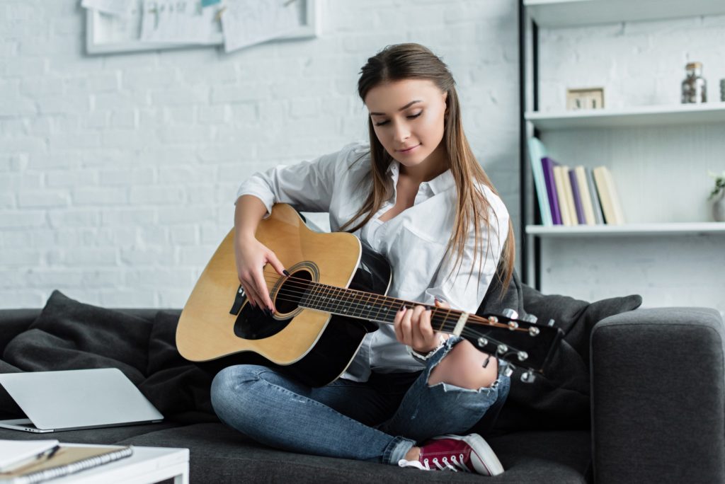 focused girl sitting on couch and playing guitar in living room