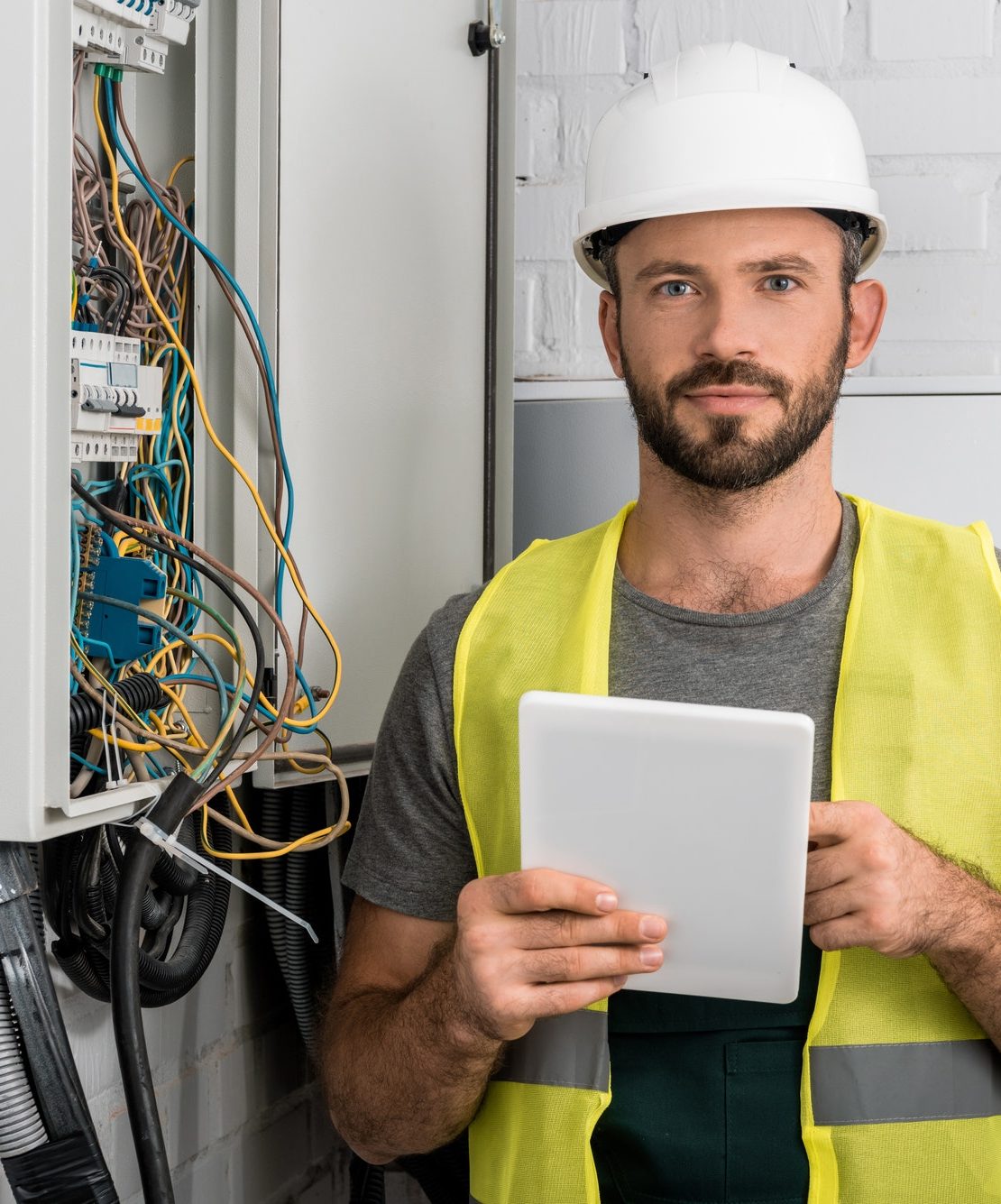 handsome electrician holding tablet near electrical box in corridor and looking at camera
