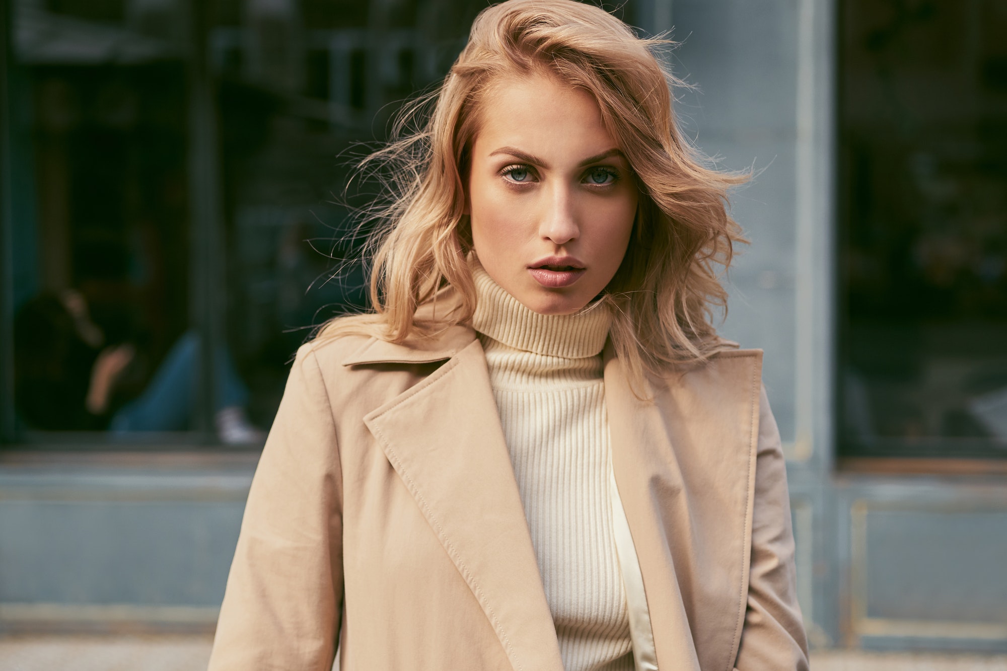 Beautiful serious blond girl in trench coat confidently looking in camera on street