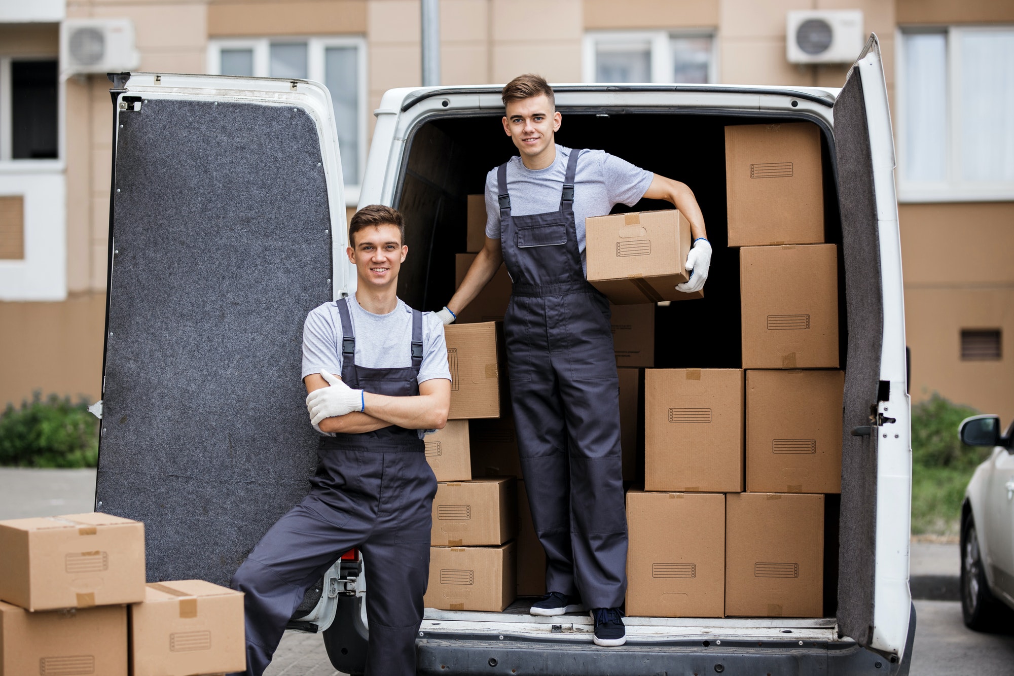 Two young handsome smiling movers wearing uniforms are unloading the van full of boxes. House move