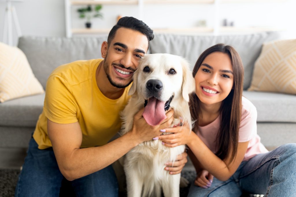 Portrait of smiling diverse couple hugging their pet dog, smiling at camera, indoors