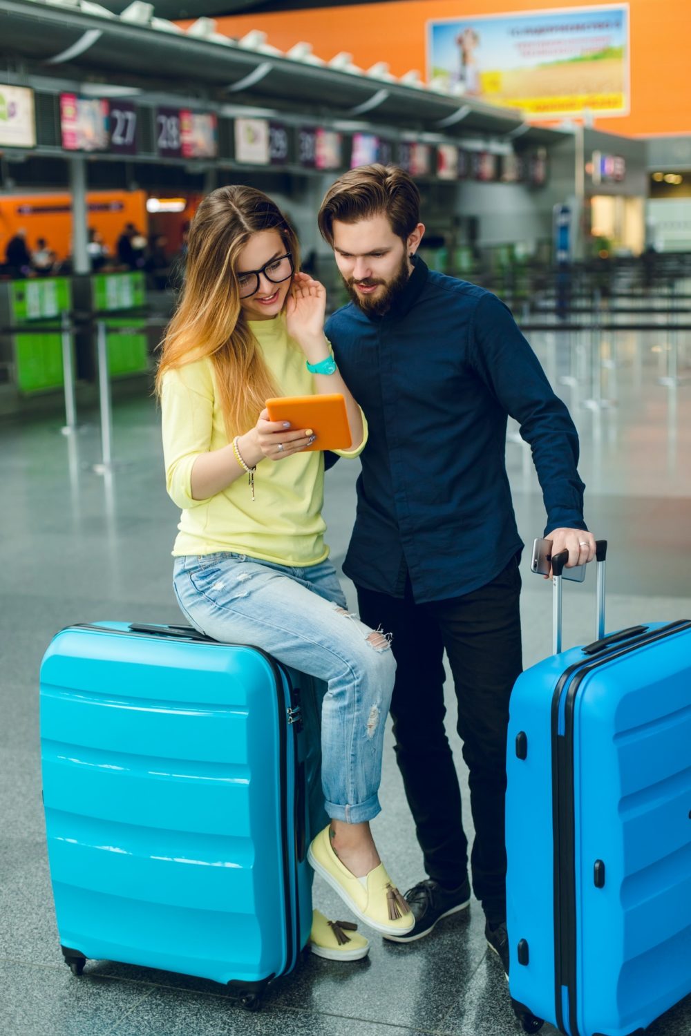 Pretty girl with long hair is sitting on suitcase in airport. Guy in shirt, pants with beard is